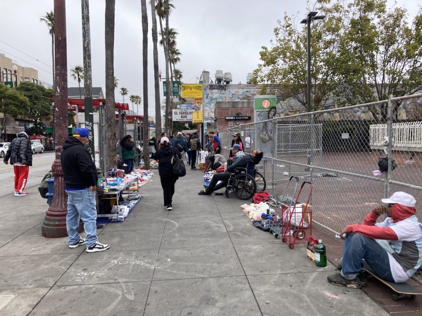 People sell goods on sidewalk outside of fenced-off plaza