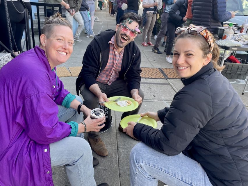 Two women and a man sitting on small stools and carrying pancakes