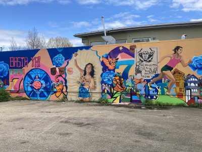 A mural with a woman raising a fist in a Virgin Mary halo, an Indigenous man pointing with the caption, "Whose the illegal alien pilgrim?" and a woman in running clothes