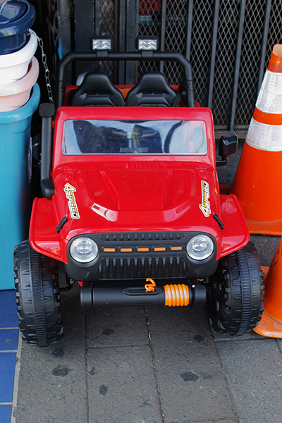 A large red toy jeep wedged between some traffic cones and some trash cans.