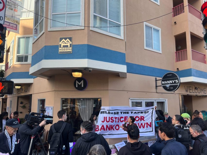 Crowd in front of a building holding a Justice for Banko Brown banner