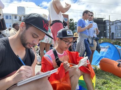 Two queer people, one wrapped in a Pride rainbow flag, sketching.