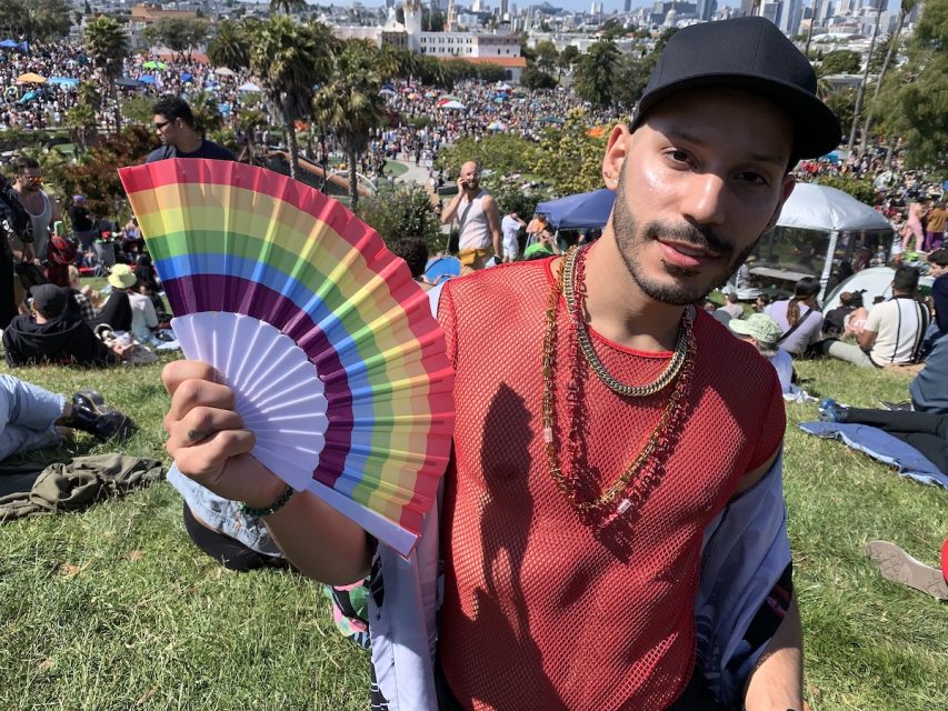 A man in a mesh red tank and two layered necklaces that read 'Pride.' He also flashes a rainbow fan.