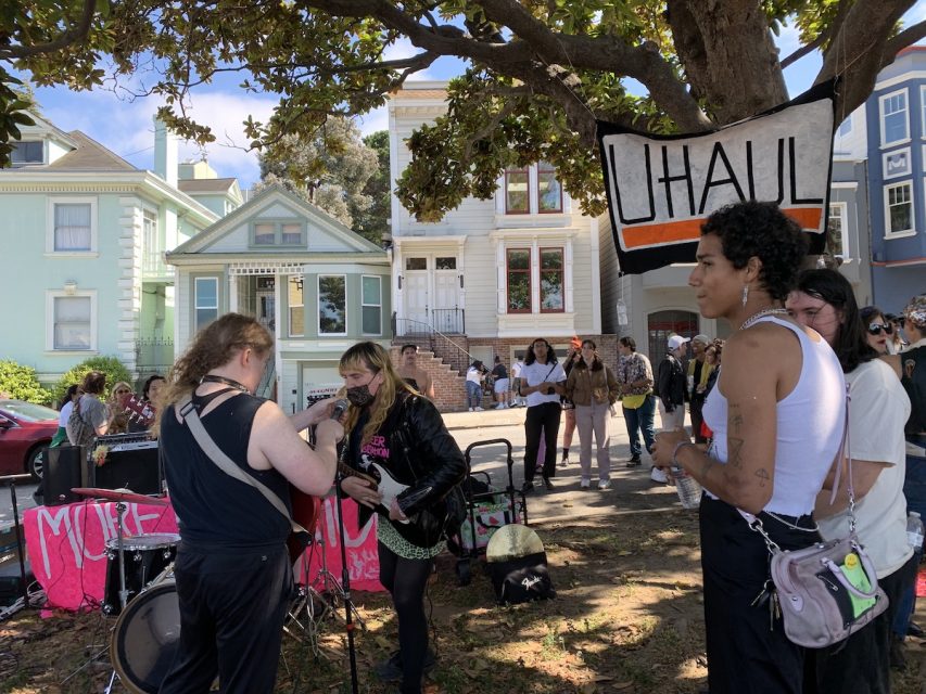 A rock bad plays behind a banner that reads, 'U-haul,' the term for lesbians partnering up and moving in together quickly.