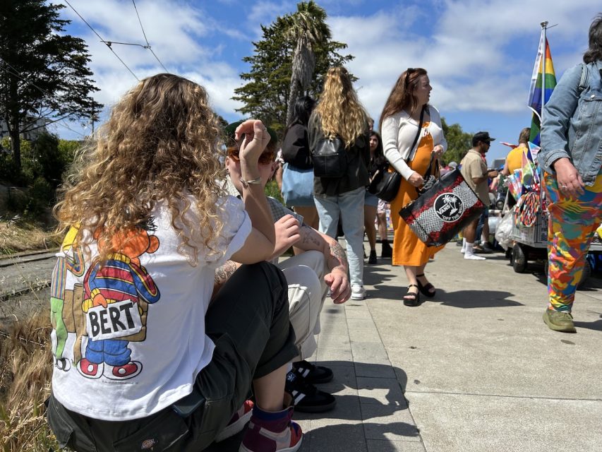 A woman with curly blonde hair wears a t-shirt depicting Sesame Street characters Bert and Ernie. At SF Pride.