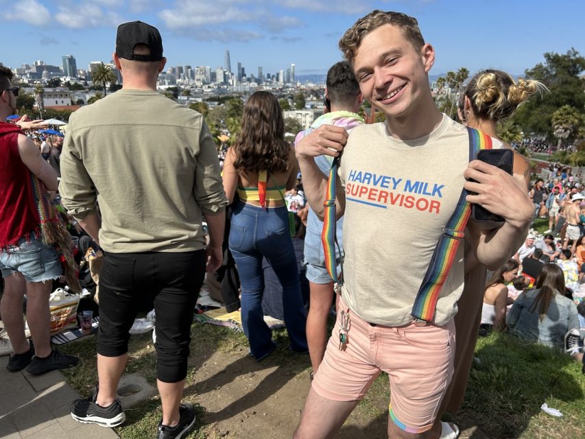 A young gay man wears a tee-shirt that says 'Harvey Milk Supervisor' and wears rainbow suspenders.