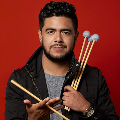 A man looking toward us with a beard and holding three drum sticks in one hand and one in another.