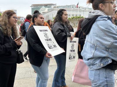 Two Latina women are holding posters that read, 'Aqui estamos, no nos vamos" outside City Hall.