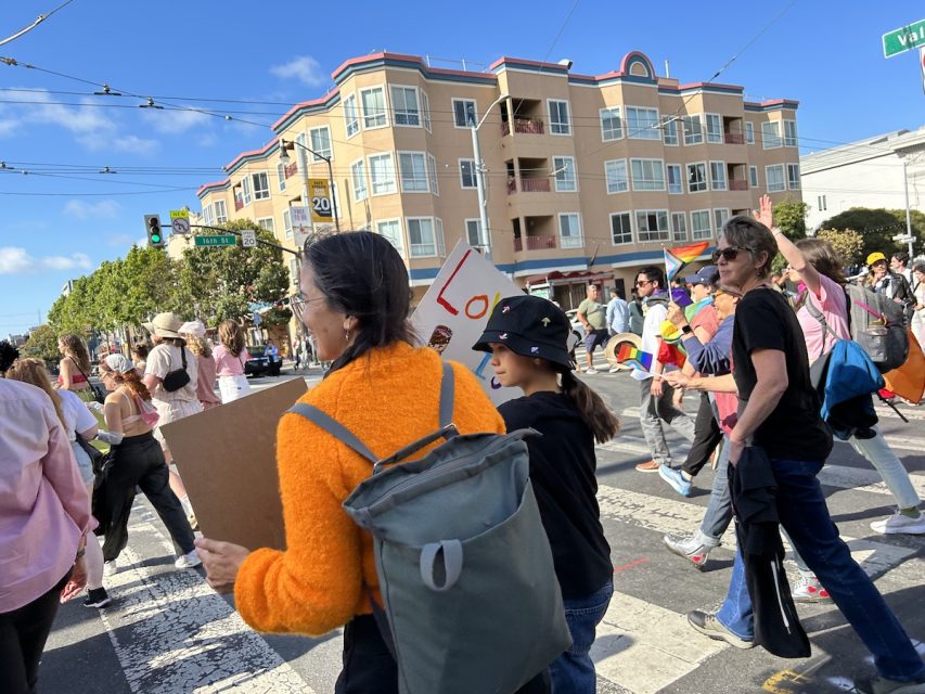 A mother in a bright orange sweater carries a sign that says 'Super Proud Mom' next to her daughter, in a bucket hat, that carries a sign that says, 'Love is Love.'