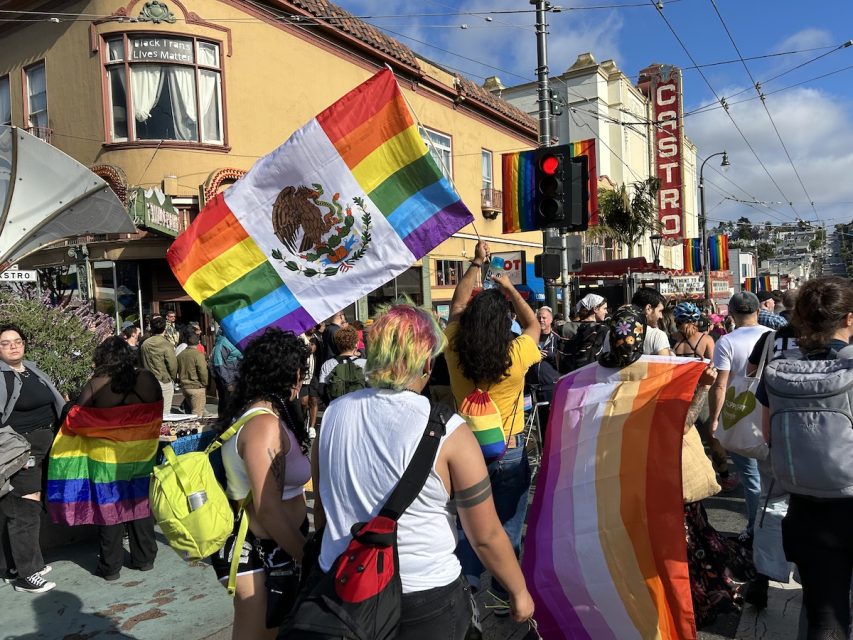 A flag is hung on the back of one lady and it has red, orange, yellow, white, light pink, magenta and burgundy stripes in that order. Another person waves a flag with a rainbow background and an eagle eating a snake, similar symbols to the Mexican flag.