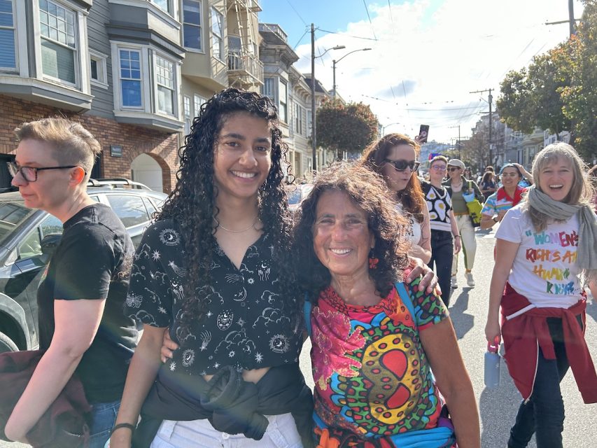 A woman in long curly hair and a celestial black crop poses with an older, sun-beaten lady with butterfly earrings and a psychadelic shirt.