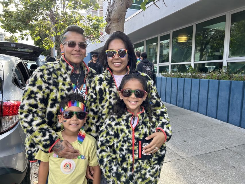 A lesbian couple and their daughter, dressed in coordinating leopard print quarter zips, and their young son dressed in a Pride bandana, pose. The parents hug the children.