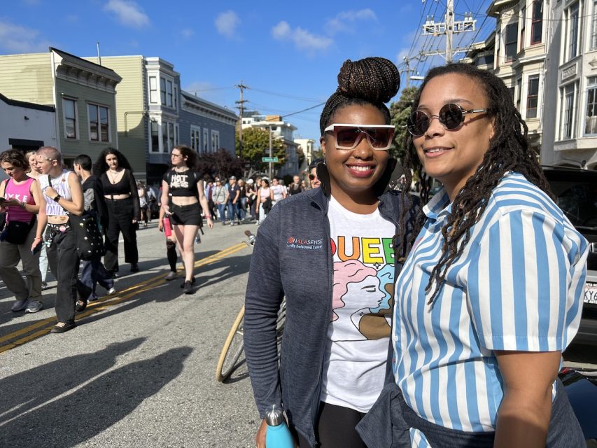 A couple, both wearing shades and Black women. One wears her hair in braids and has a blue and white striped shirt. The other, holding her around the waist, wears a tee that reads 'Queer' and depicts two women.