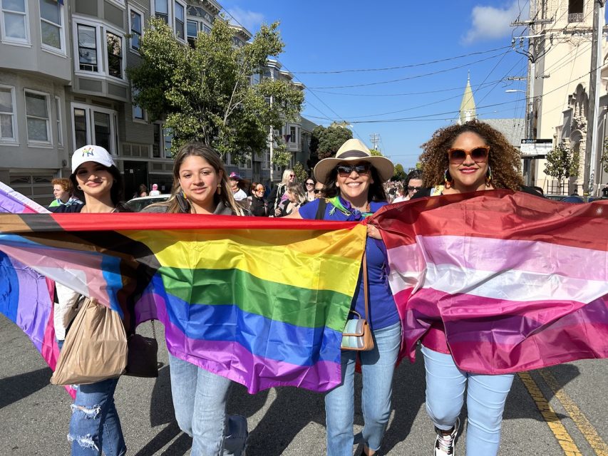 Four Latina women, two adolescents, carry multiple Pride flags. 