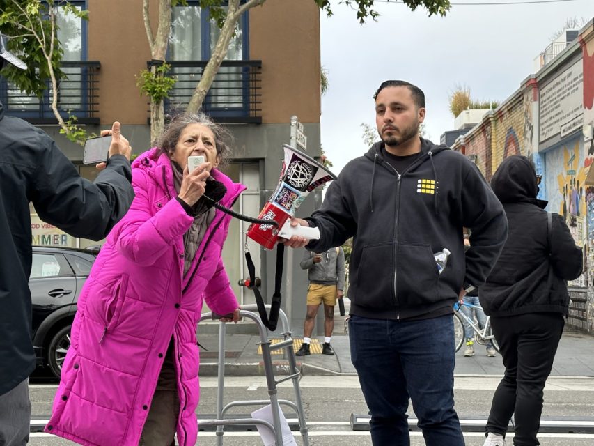 woman in pink coat speaking into bullhorn held by mad in black jacket