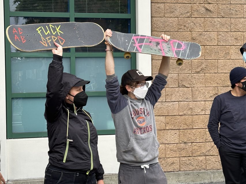 two skateboarders and protesters holding skateboards reading "Fuck SFPD" and "FTP"