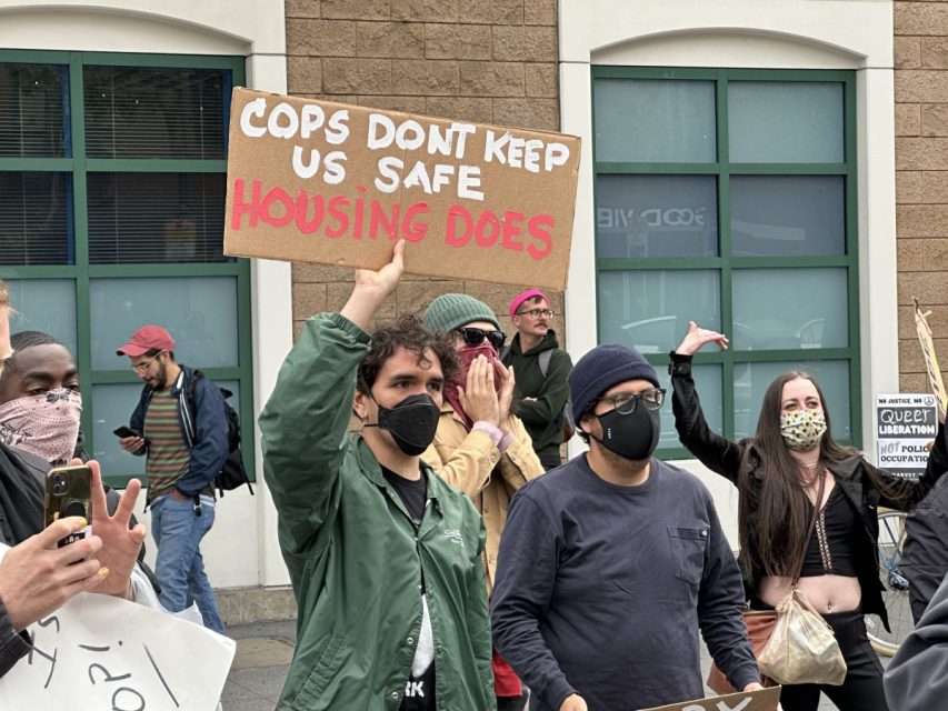 Masked protester holding a "cops don't keep us safe, housing does" sign