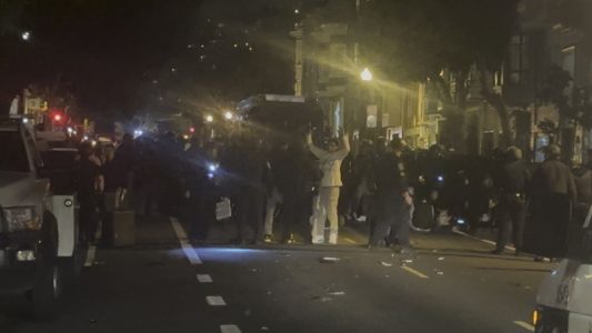 A night shot of teenagers with hands up, officers, vans after the 2023 Dolores Park hill bomb