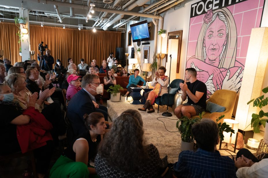 Two people onstage in a crowded room, a large poster of a figure with many hands behind the two seated speakers.