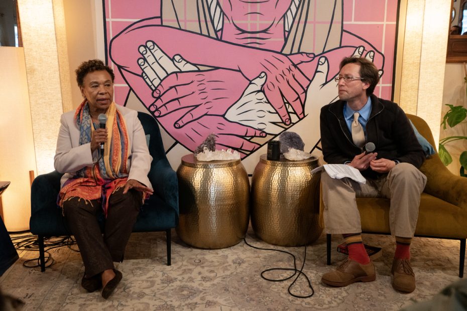 A Black woman and a white man with glasses seated on a stage with gold side tables and a mural of pink and white hands behind them.