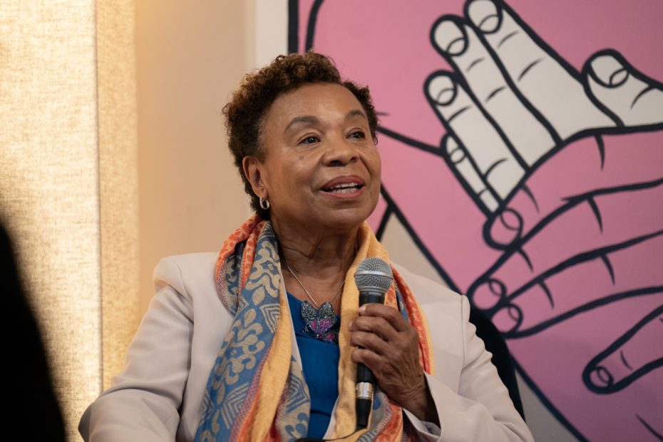A Black woman with an orange scarf standing in front of a. mural with pink and white hands.