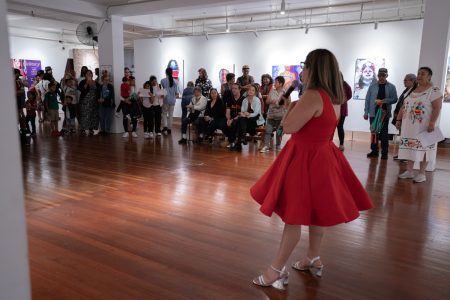 A crowd at a gallery and a woman in a red dress talking to them.