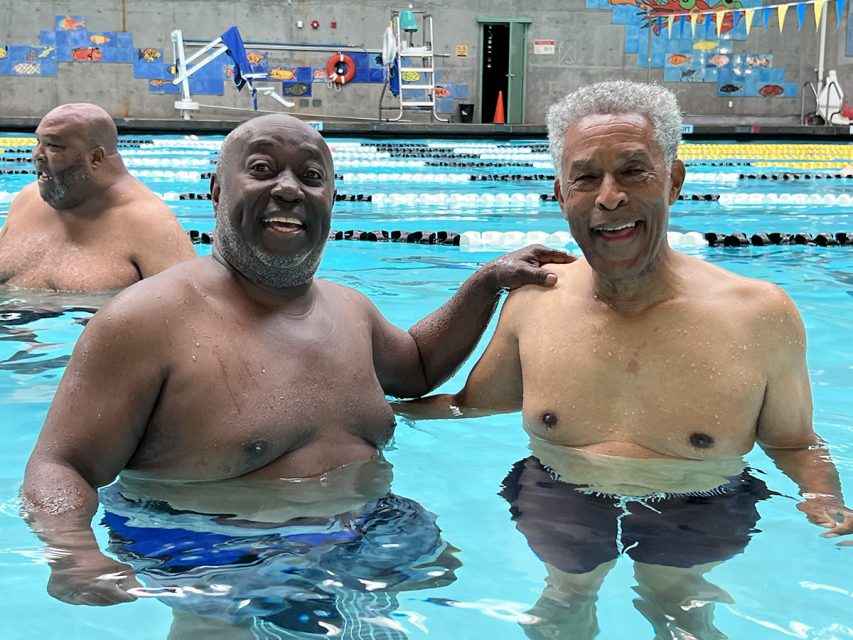 Two older men one with his arm on the other, facing forward in a swimming pool.