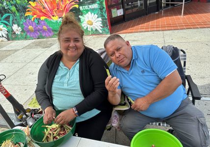 Two people, one with a black sweater and blue shirt and the other, a man in a blue shirt. They are seated and shelling beans.