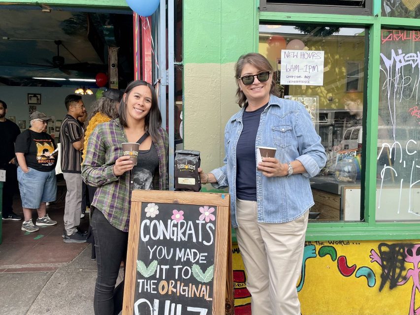 Two women standing in front of a Philz coffee shop with a sign.
