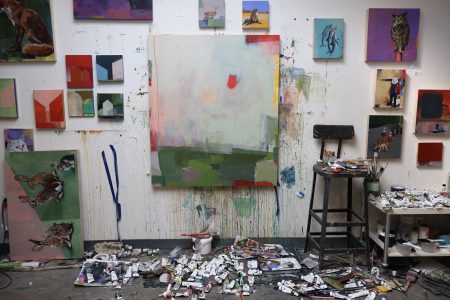 An artist's studio with a lot of paintings on the wall and paints scattered on the floor.