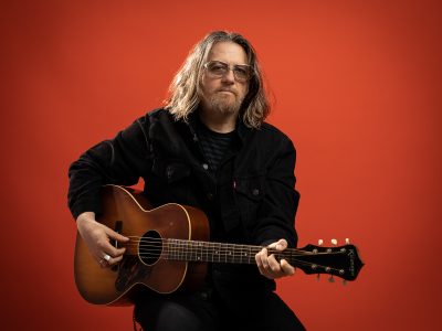 Tom Heyman holding an acoustic guitar in front of a red background.