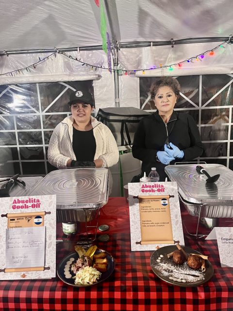 Two women standing in front of a tent with food they made on the table. 