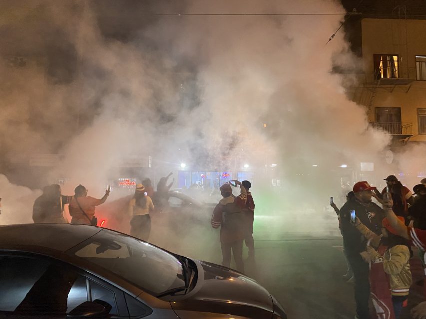 A group of people standing in front of a car with fireworks smoke around it, celebrating the 49ers win