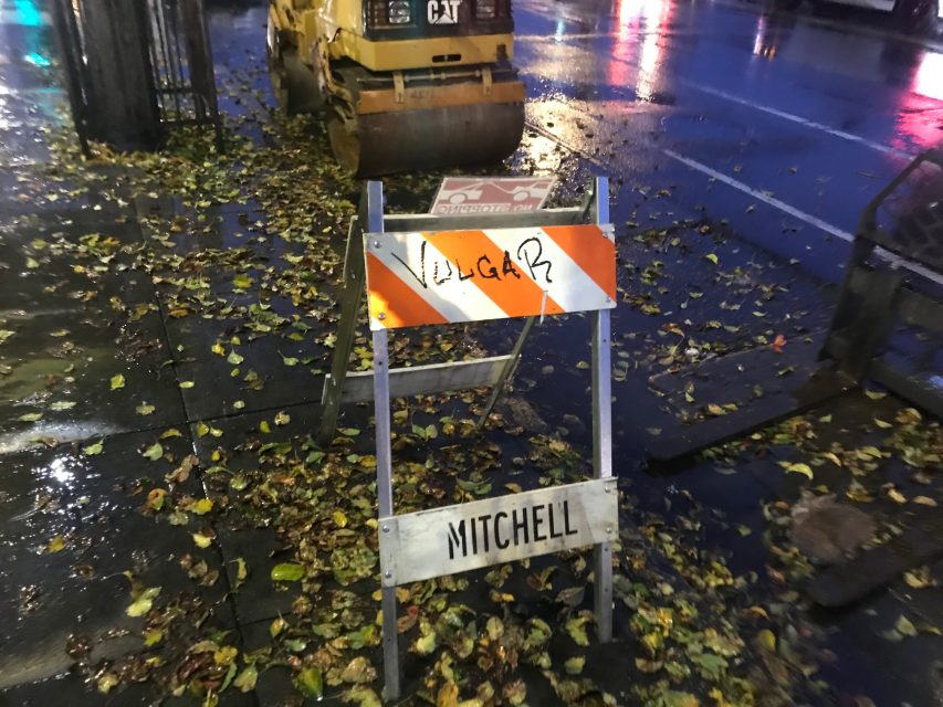 A traffic barricade with the words 'Vulgar' and 'Mitchell' on the wet sidewalk.