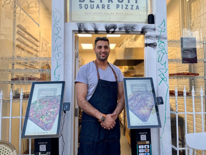 A man in an apron standing in front of a pizza shop.