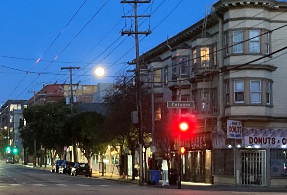 City street at dusk with a full moon rising above, traffic light on red, and illuminated storefront signs.