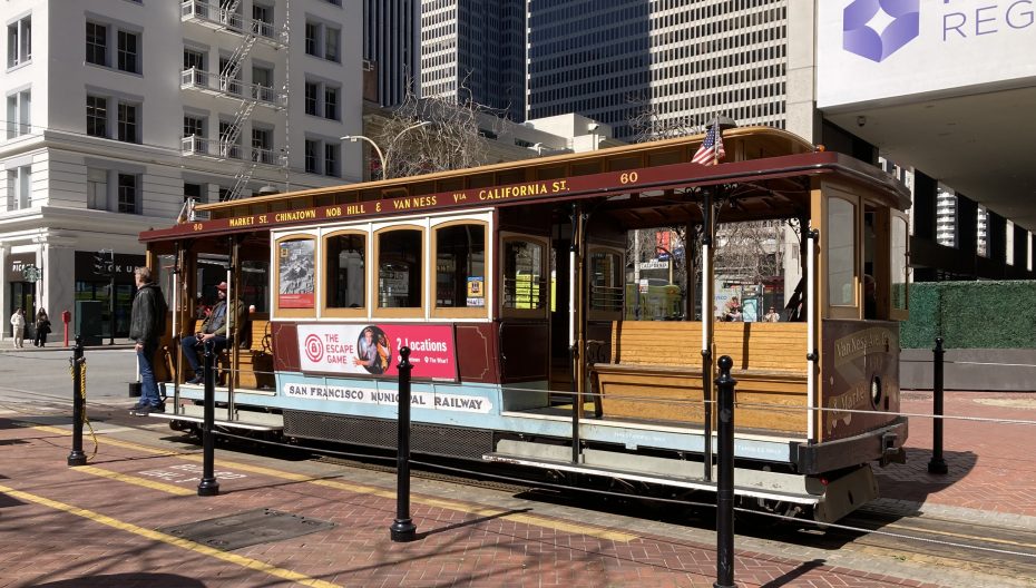A cable car at a stop in san francisco, with passengers on board and surrounding buildings in the backdrop.