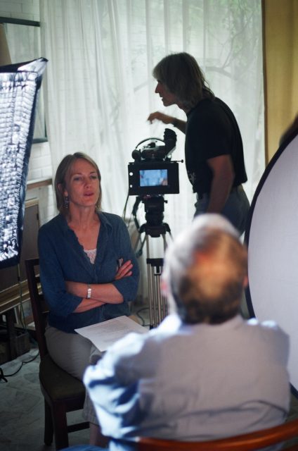 Behind-the-scenes view of a video interview setup with a seated female subject, camera operator, and lighting equipment.