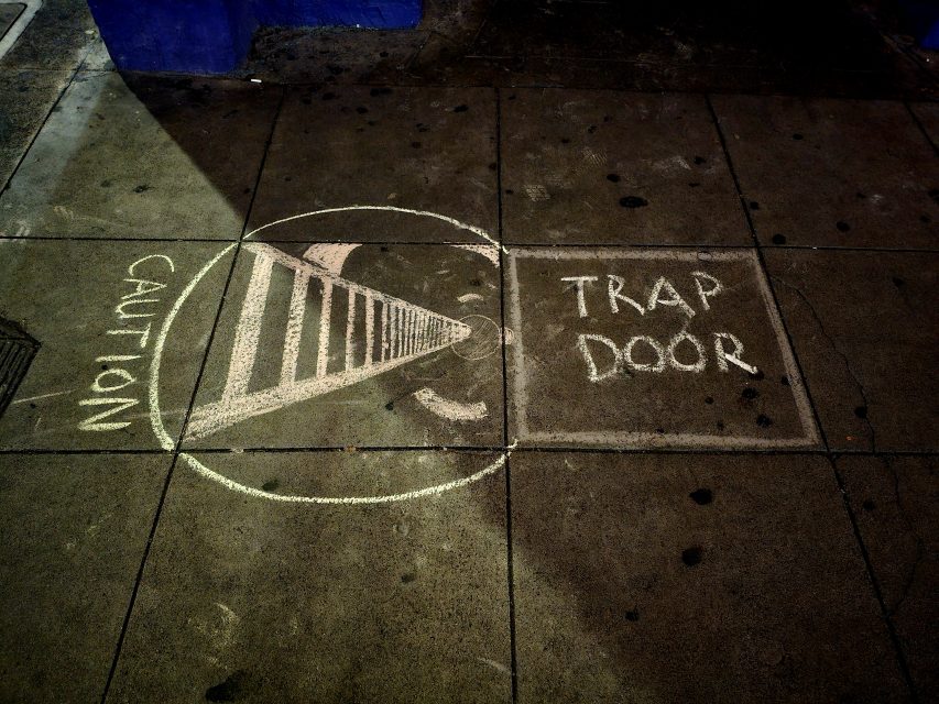 Chalk drawing on pavement depicting a cone with "caution" written above and "trap door" written beside, implying a hidden hazard.