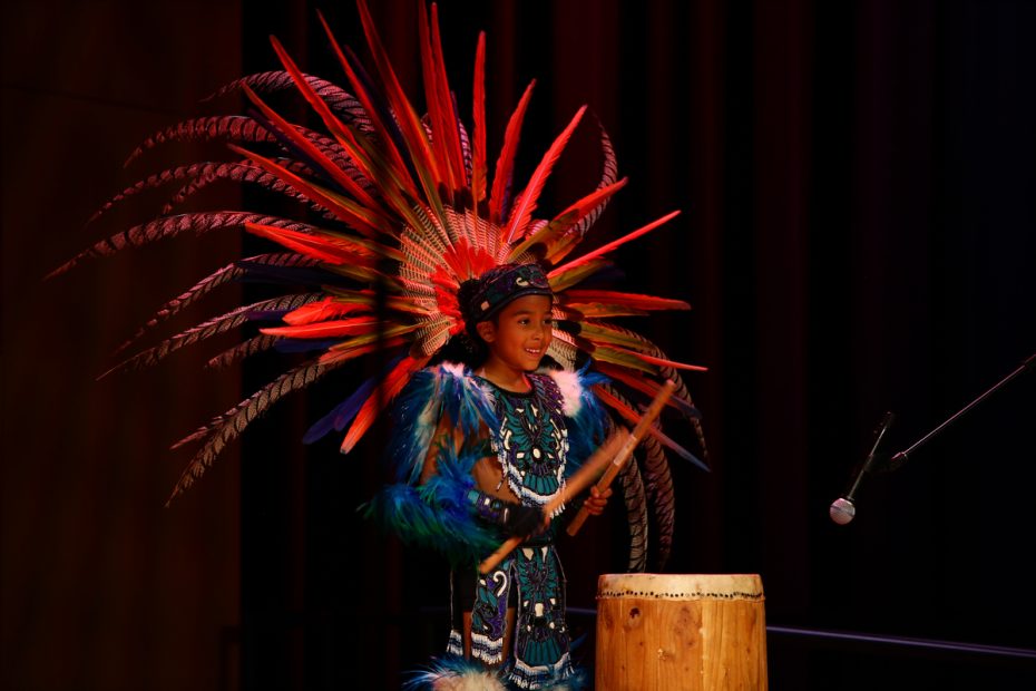 A child in vibrant traditional attire with feathered headdress playing drums on stage.