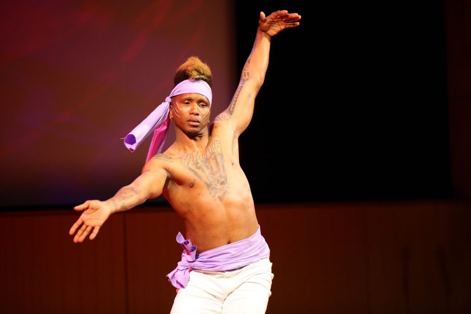 A shirtless performer with a purple headband and wrap gestures expressively on stage.