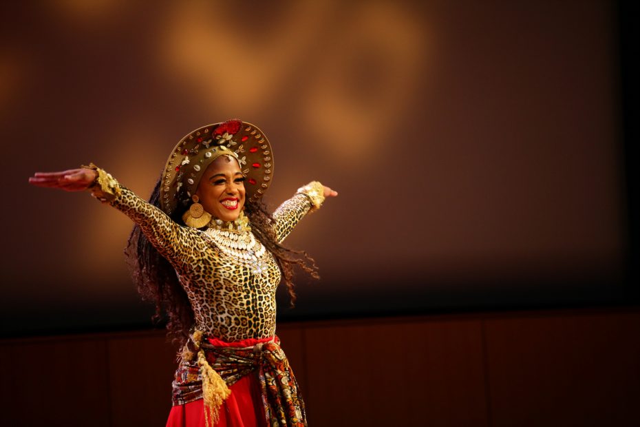 A performer in vibrant, traditional costume smiling widely while dancing on stage.