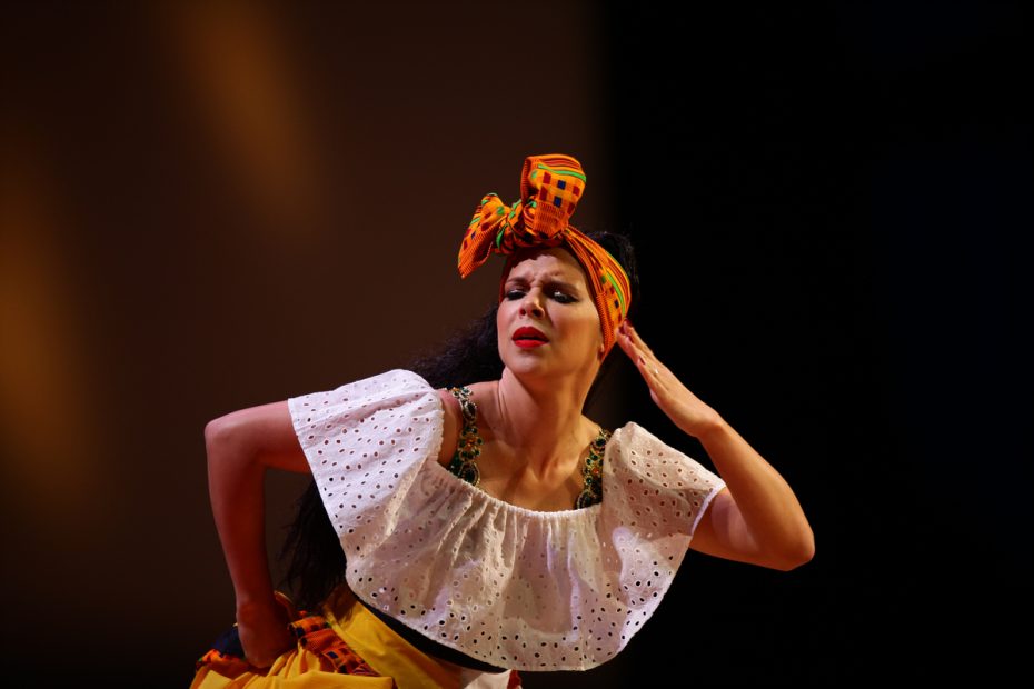 A dancer in colorful costume performing with a dramatic expression under stage lighting.