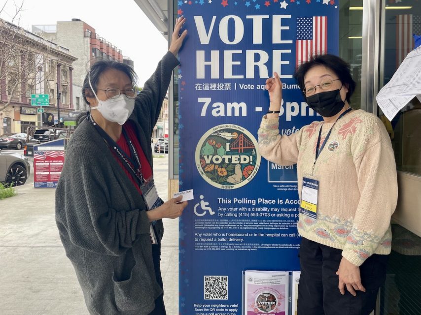 Two individuals wearing masks are standing next to a "vote here" sign, pointing at it and displaying their "i voted!" stickers, indicating they have participated in an election.