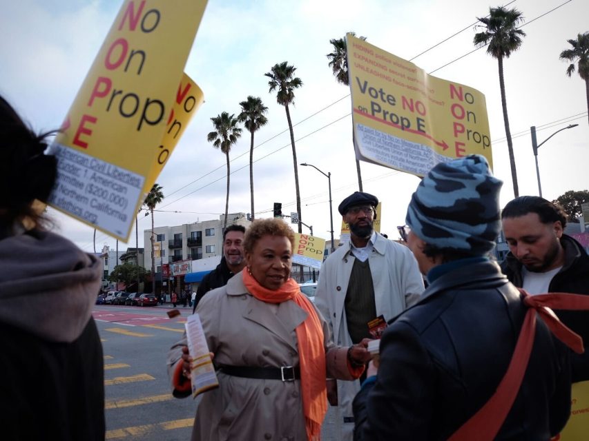 Activists gathered around Barbara Lee at the 24th St. BART plaza, holding "No on Prop. E" signs