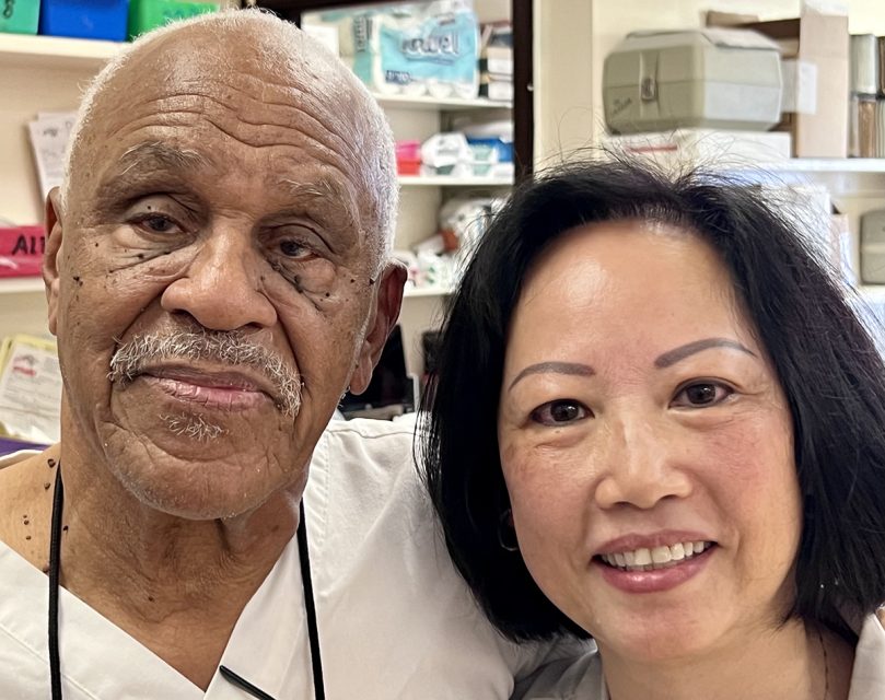 Two smiling individuals, one elderly man with a mustache and one middle-aged woman, posing together indoors, possibly in a workplace setting.