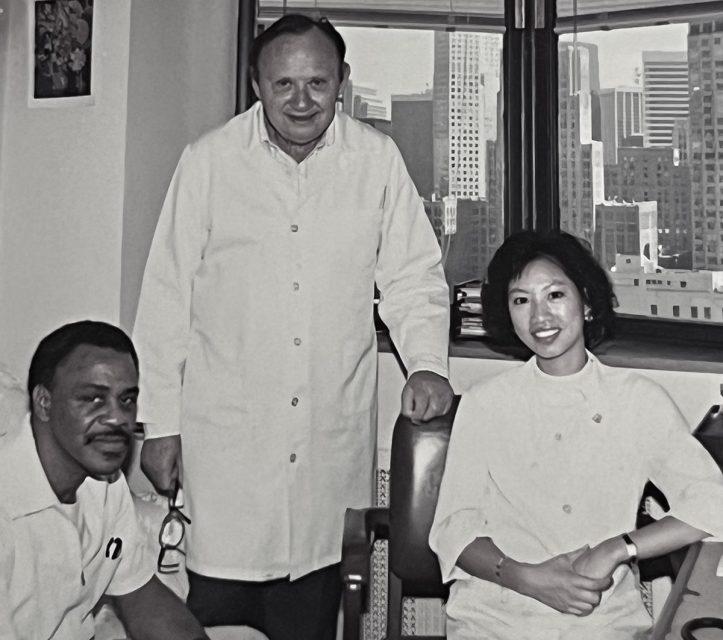 Three healthcare professionals posing for a photo in a clinic with a cityscape visible through the window in the background.