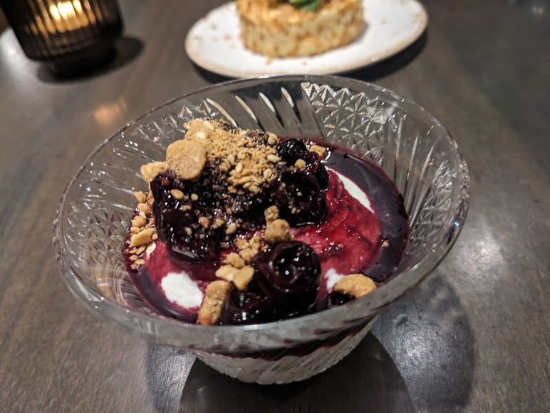 A close-up of a berry-topped dessert with crumbled nuts in a glass bowl, served on a dark wooden table.