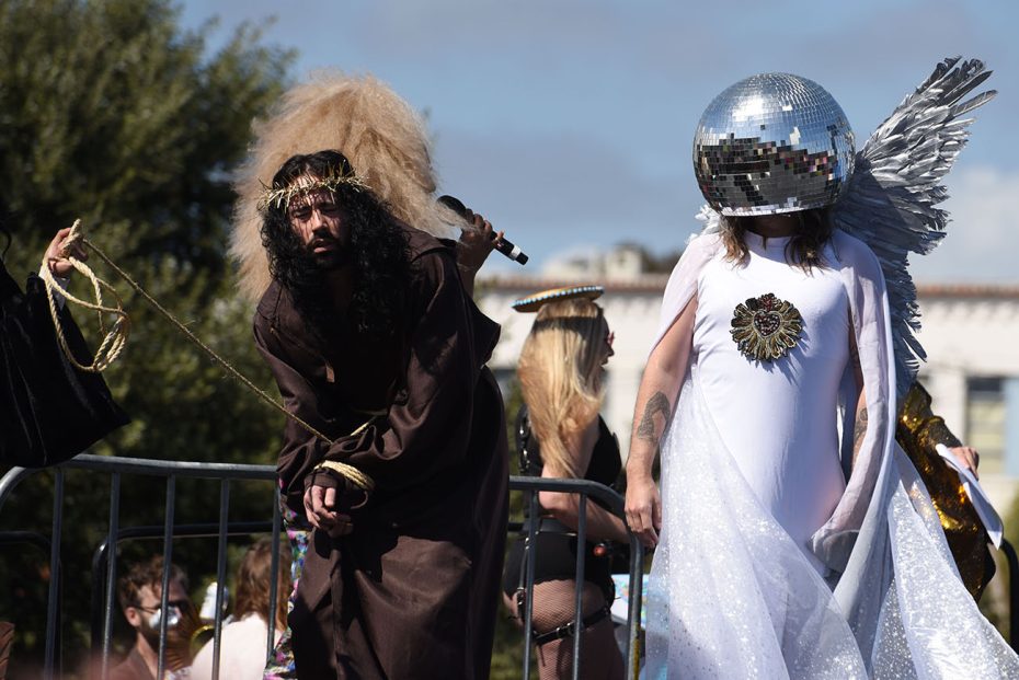 Two individuals dressed in costume, one as a rugged character with a whip, the other wearing a mirrored disco ball headpiece and wings, participating in Hunky Jesus.