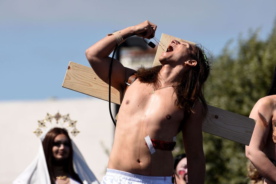 A person performing a dramatic enactment, possibly of a religious or historical scene, with intense expression and a mock crucifixion setup at Hunky Jesus.